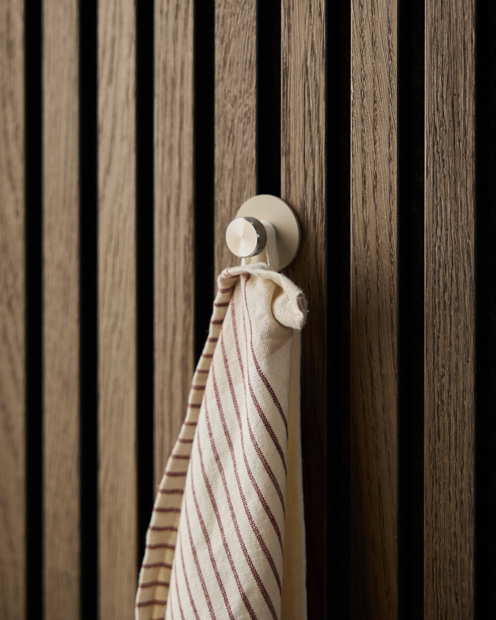 Pasiasty ręcznik wisi na wieszaku WoodUpp beige Norr Coat Hook, wykonanym ze stali nierdzewnej steel, zamontowanym na teksturowanym drewnianym panelu akustycznym z lamelami Akupanel.