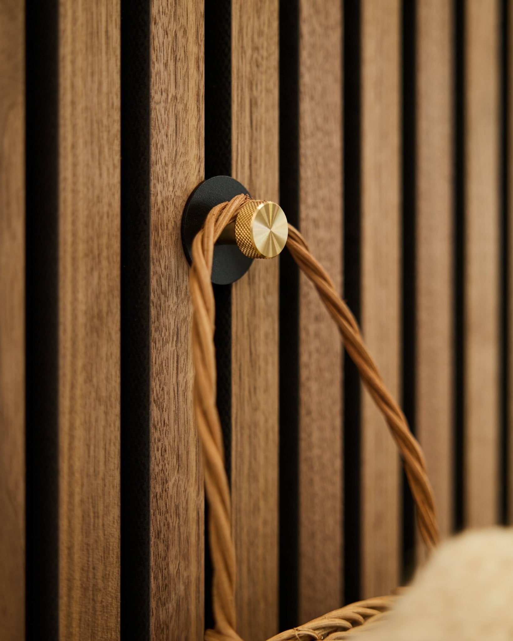 Wieszak na płaszcz WoodUpp Norr w kolorach black i brass, zamontowany na drewnianej podstawie Akupanel Panel akustyczny, podtrzymuje pleciony uchwyt w jasnobrązowym odcieniu.