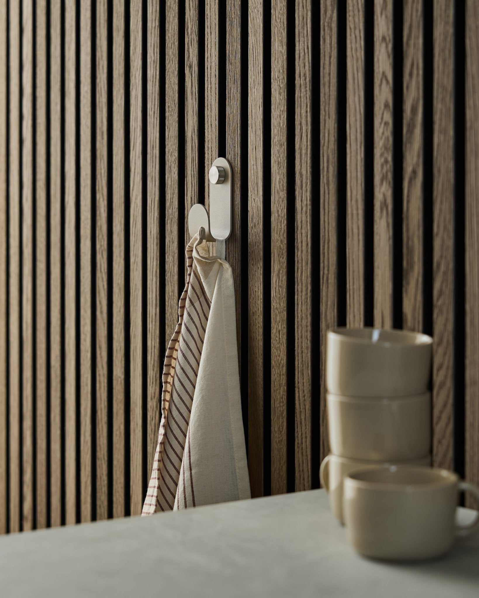 Pasiasty ręcznik wisi na WoodUpp Norr Single Hanger w kolorze beige, zamontowanym na akustycznym panelu lamelowym w stylu nordyckim; z przodu ustawiono trzy kubki beige.