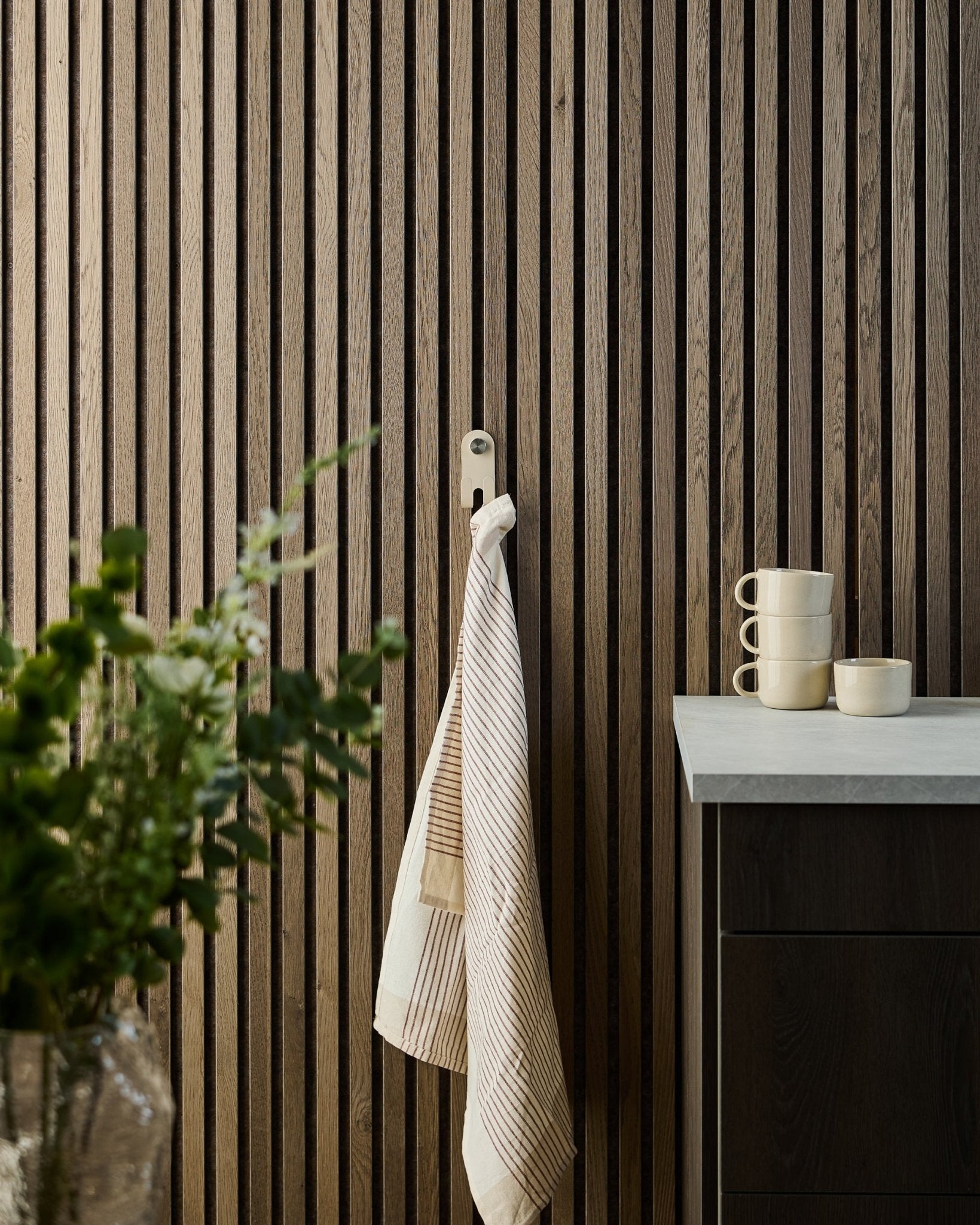 Pasiasty ręcznik wisi na WoodUpp Norr Double Hanger w kolorze beige, zamontowanym na nordyckim akustycznym panelu z listew; poniżej stoją kubki, miska i zielona roślina.
