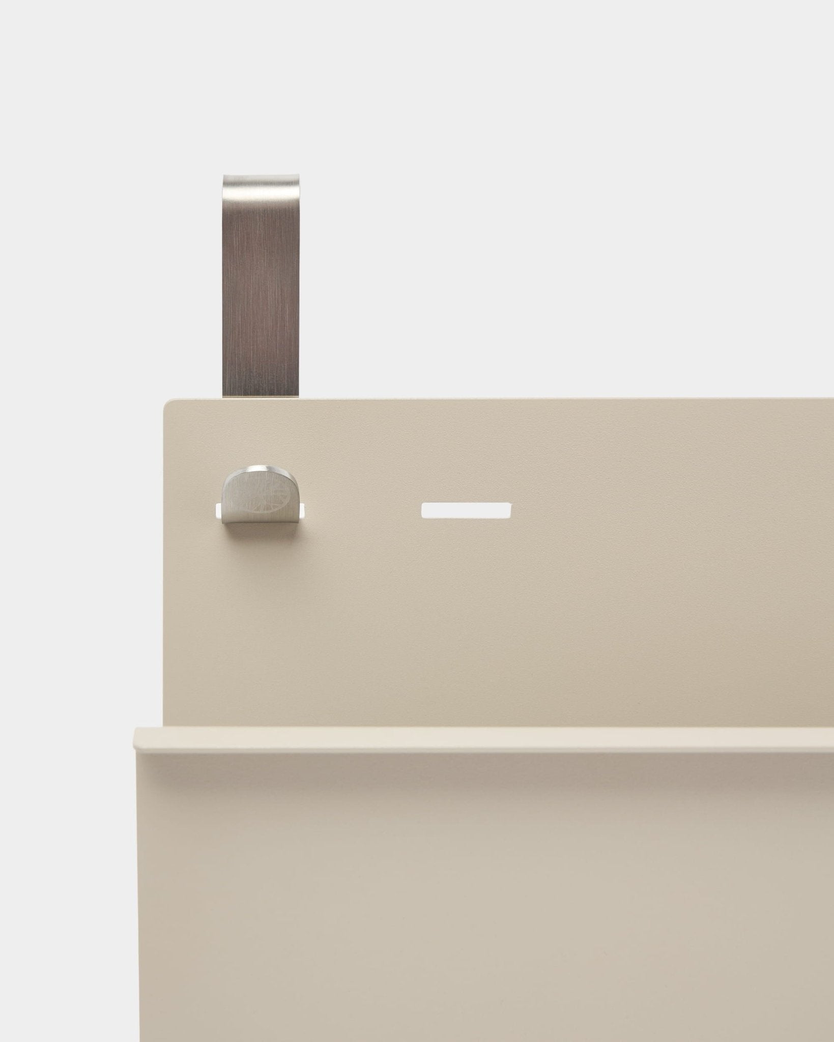 Zbliżenie na Nell Magazine Storage od WoodUpp z malowanego proszkowo steel w kolorze beige, z półką, szczeliną, śrubą i zaokrąglonym uchwytem; pełni funkcję ścianki działowej lub uchwytu na czasopisma.