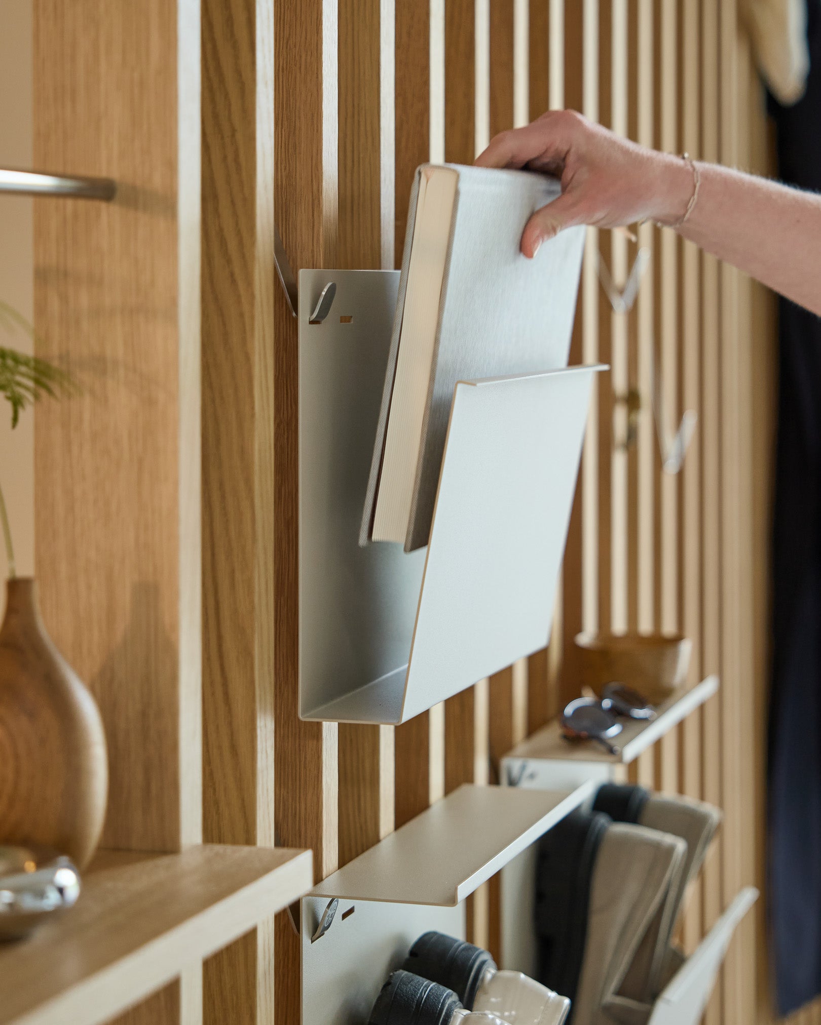 Dłoń odkłada Nell | Magazine Storage / Beige od WoodUpp na malowaną proszkowo półkę steel, zamontowaną na nordyckim drewnianym Panel akustyczny, obok innych przedmiotów.