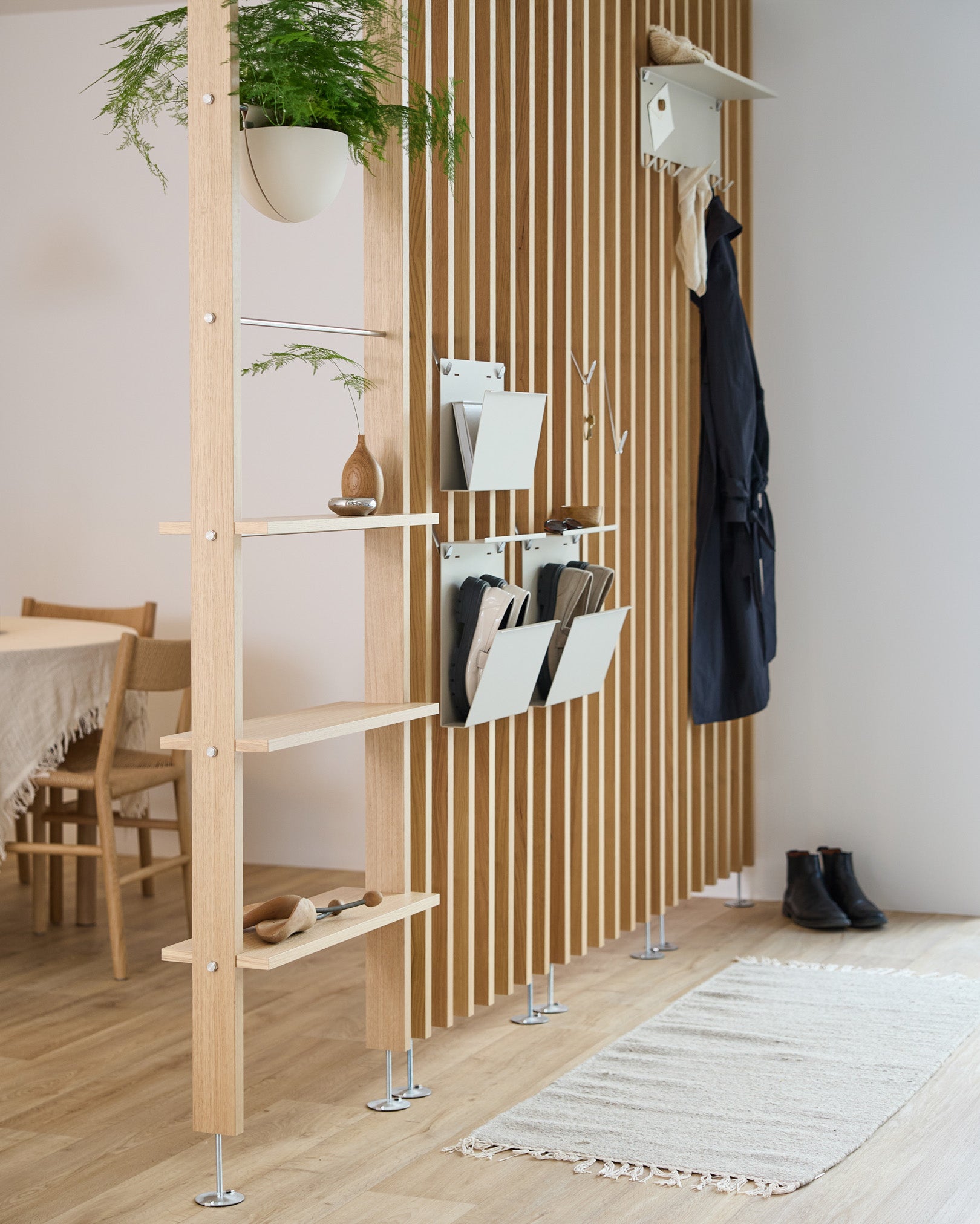 Nell Magazine Storage od WoodUpp w kolorze beige, zintegrowany ze ścianką działową z półkami steel, pojemnikami do przechowywania i haczykami na płaszcze; w tle widać jadalnię i dywan.
