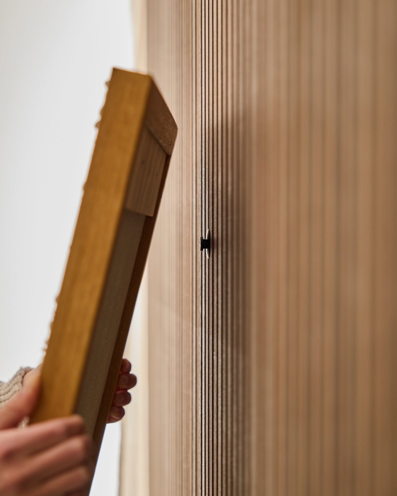 Osoba używa zestawu WoodUpp Dupps | Original do montażu ramy Akupanel akustycznego panelu drewnianego, skupiając się na przymocowaniu haka small black w pobliżu lameli vertical.
