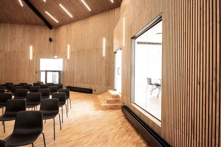 Przestronna sala konferencyjna z drewnianymi panelami akustycznymi Akupanel na ścianach i suficie, rzędami czarnych krzeseł, dużymi oknami i pionowymi oprawami oświetleniowymi.