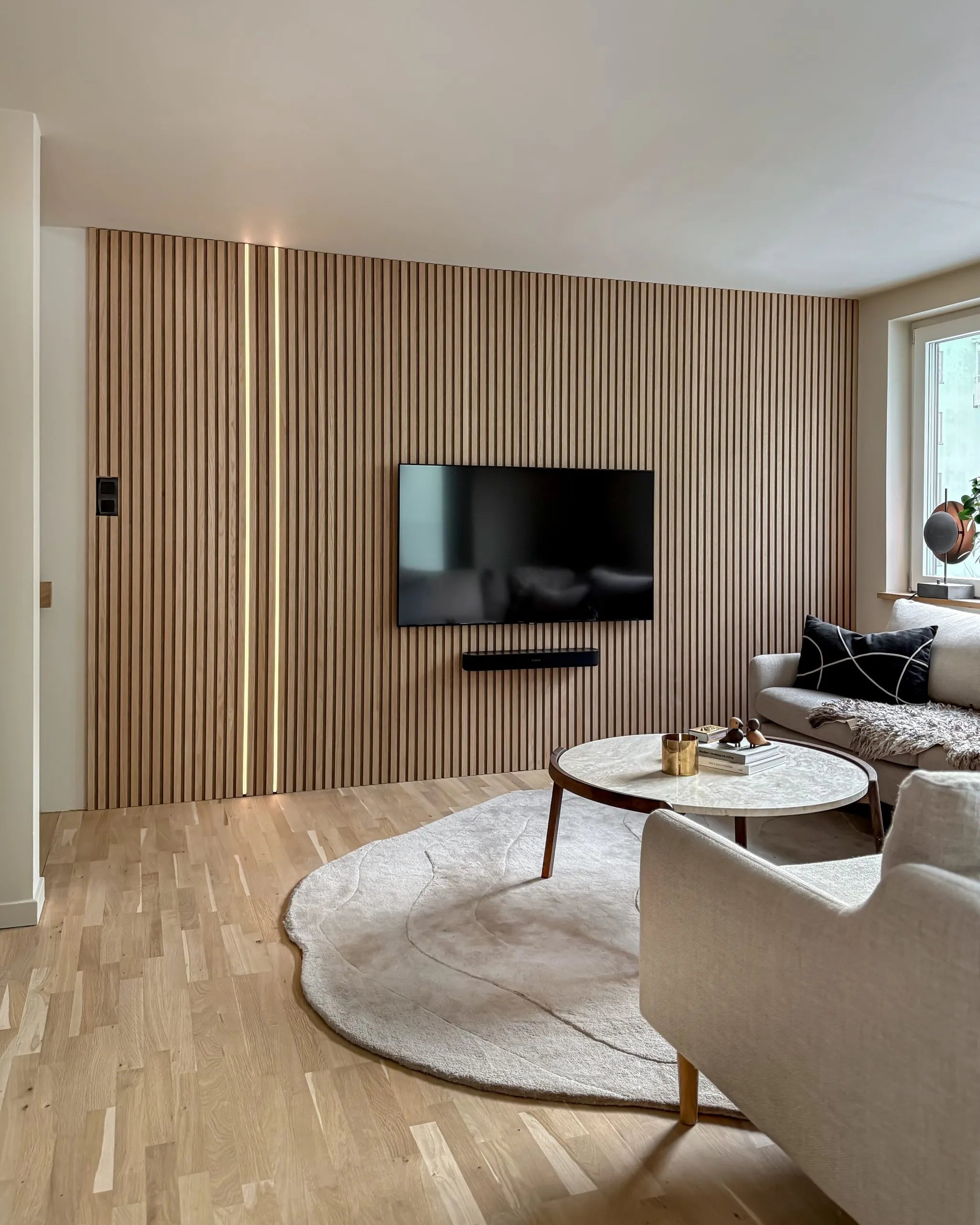 Nowoczesny salon ze skandynawską ścianą z drewnianych listew akustycznych Akupanel, zamontowanym telewizorem, okrągłym stolikiem kawowym, kremową sofą, dużym dywanem na jasnej drewnianej podłodze.