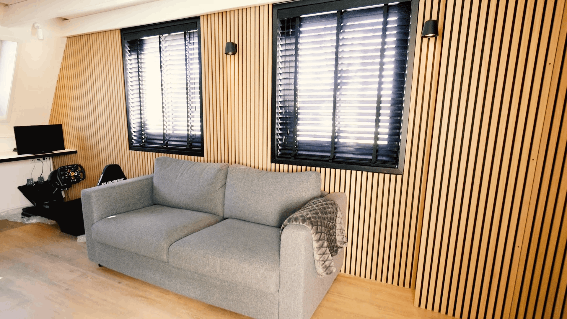 Szara sofa z wzorzystym kocem umieszczona przed skandynawską ścianą z drewnianych listew akustycznych Akupanel, otoczona dwoma oknami z czarnymi roletami i kompaktowym biurkiem.