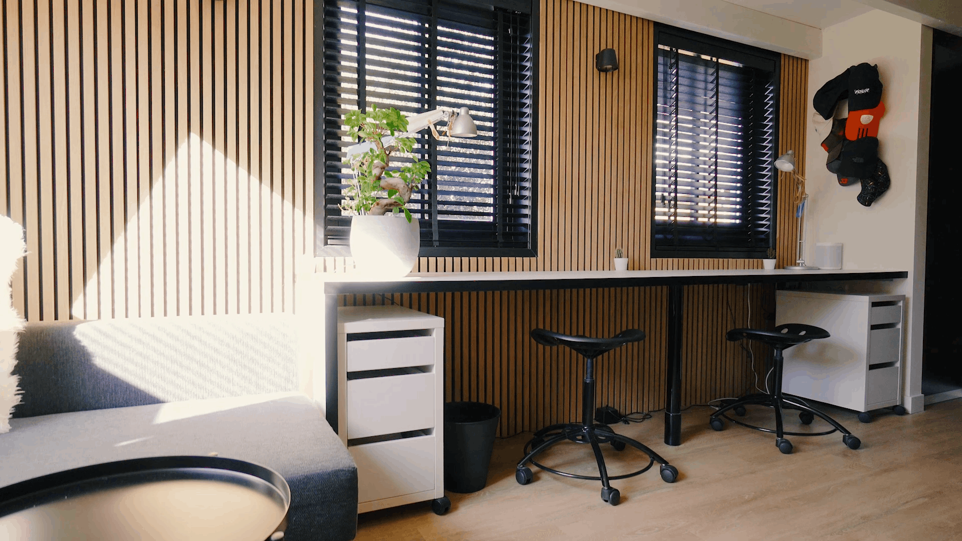 Nowoczesne biuro z nordyckimi panelami akustycznymi na ścianach, ergonomicznymi stanowiskami pracy, czarnymi stołkami, rośliną doniczkową i światłem słonecznym przez czarne rolety okienne.