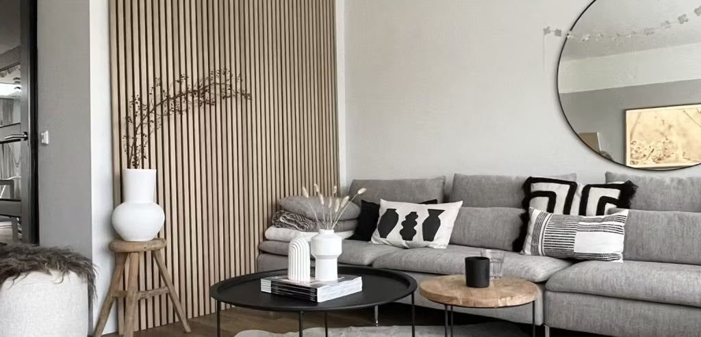 Nowoczesny salon z szarą sofą, abstrakcyjnymi poduszkami, czarnymi stolikami kawowymi, dekoracyjnymi wazonami i akustyczną ścianą z drewna Akupanel z okrągłym lustrem.