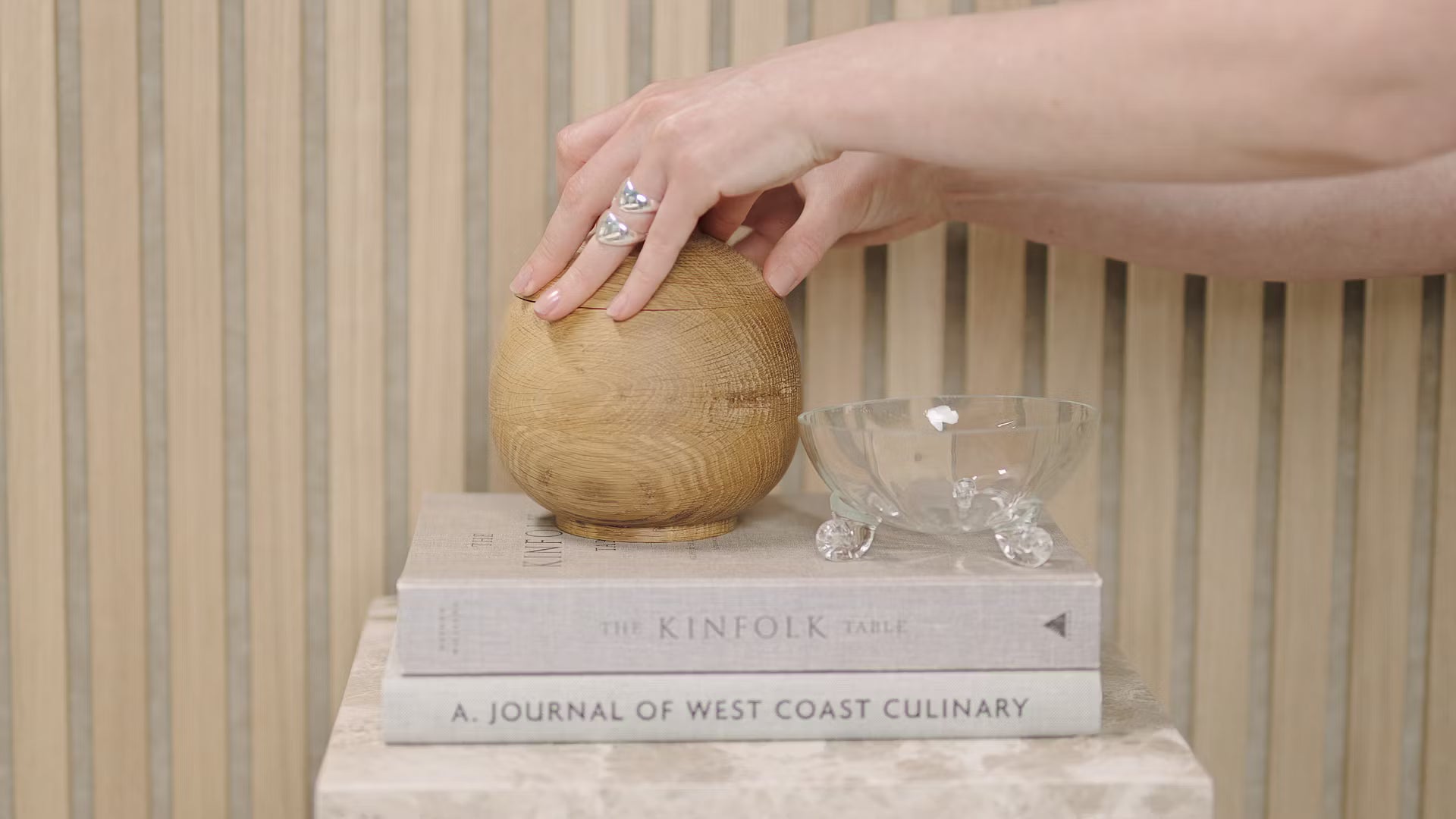 Dłoń dotyka drewnianej kuli umieszczonej na ułożonych w stos książkach, w tym Kinfolk Table i Journal of West Coast Culinary, obok szklanej miski na panelu akustycznym z drewnianych listew.