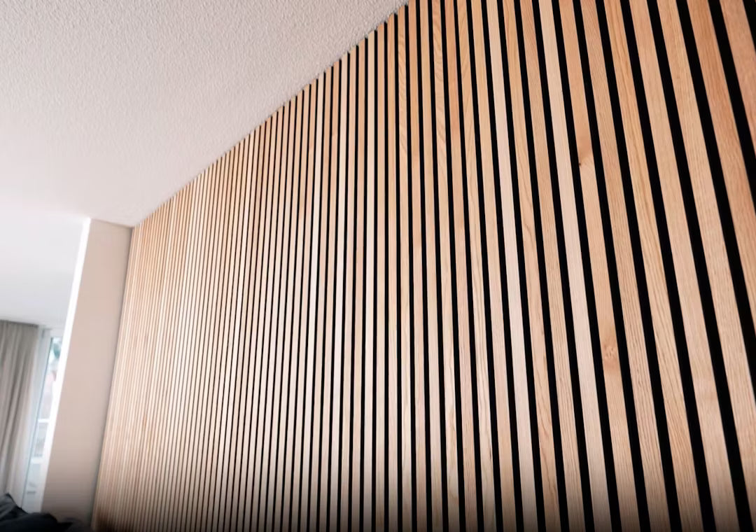 Zbliżenie drewnianego panelu akustycznego Akupanel z pionowymi drewnianymi listwami w paski, zainstalowanego na wewnętrznej ścianie pod białym sufitem.