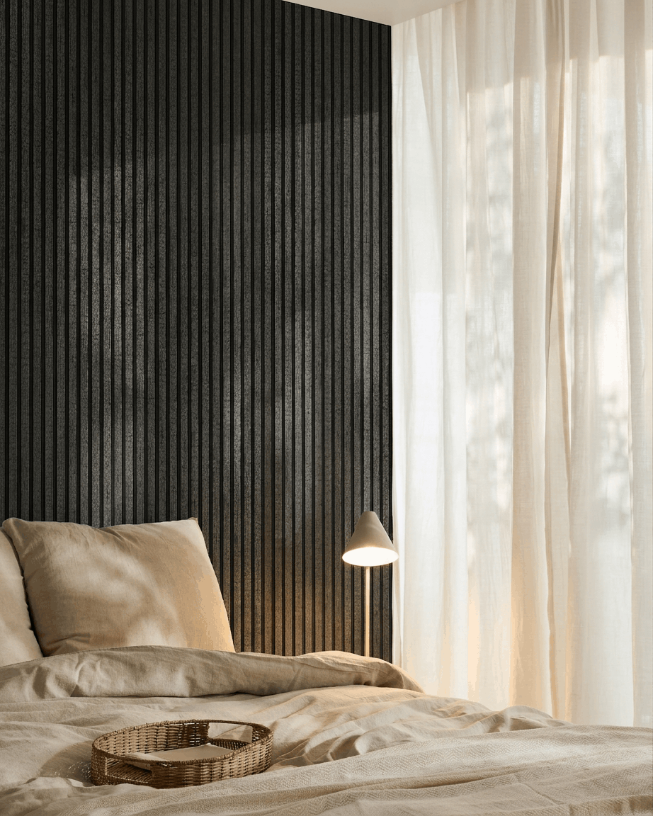 Przytulna sypialnia z pościelą beige, rattanową tacą, białą lampą, lekkimi zasłonami i akustycznym panelem drewnianym WoodUpp Akupanel w kolorze charcoal zamontowanym na ścianie.