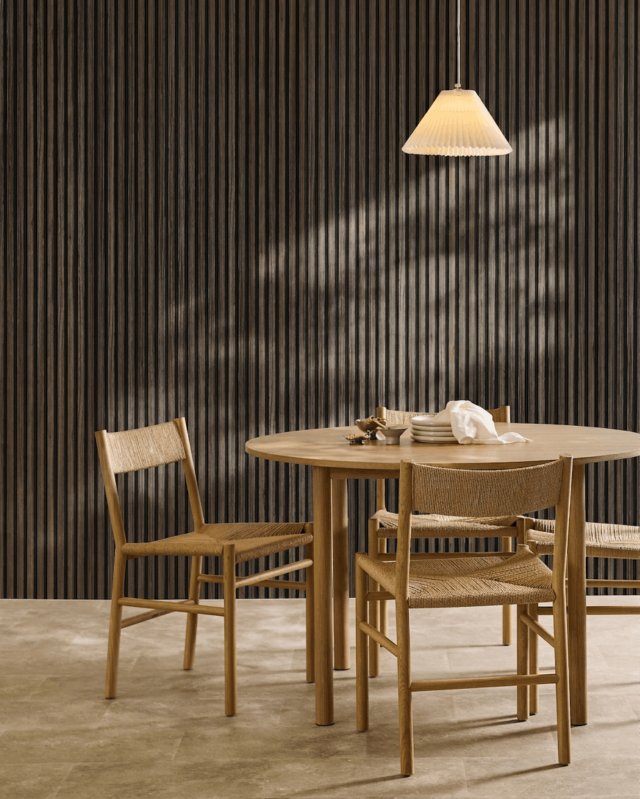 Okrągły stół z czterema plecionymi krzesłami ustawiono obok panelu WoodUpp Akupanel Black Oak Panel akustyczny, pod plisowaną lampą wiszącą.