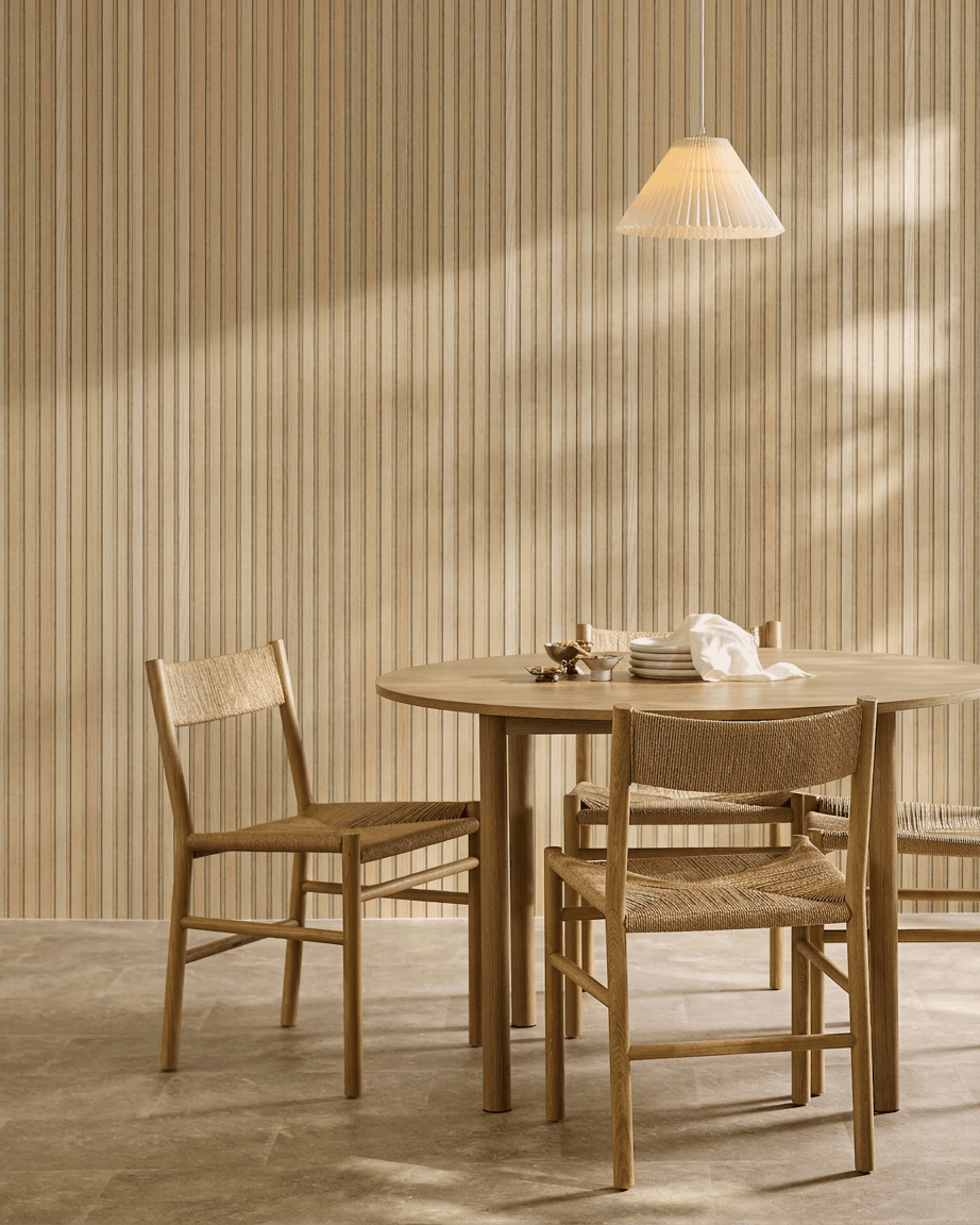 Minimalistyczna jadalnia z okrągłym stołem, plecionymi krzesłami i lampą wiszącą obok akustycznego panelu drewnianego WoodUpp Akupanel w kolorze Classic Oak z filcem beige.