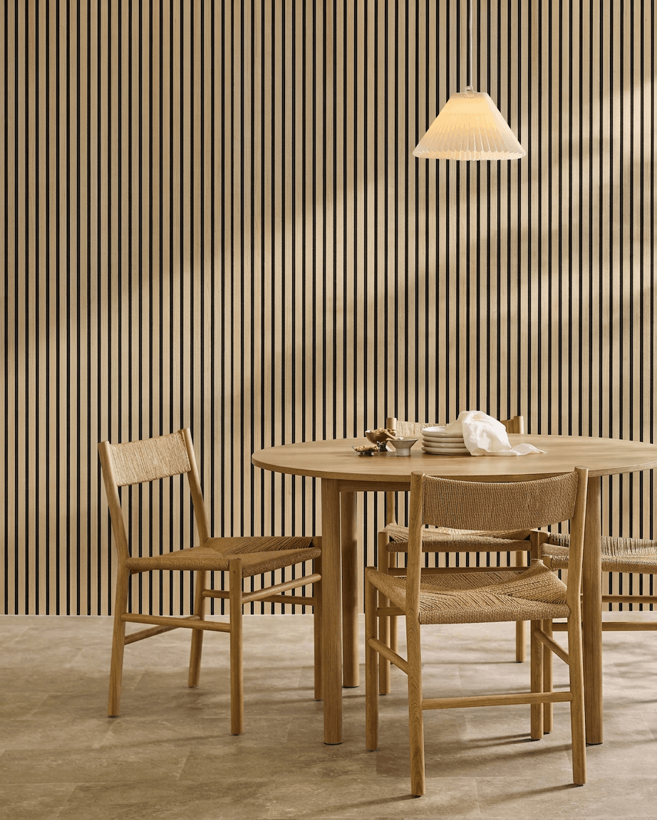 Minimalistyczna jadalnia z okrągłym stołem, plecionymi krzesłami, lampą wiszącą i panelem WoodUpp Akupanel Classic Oak Panel akustyczny zamontowanym na ścianie.