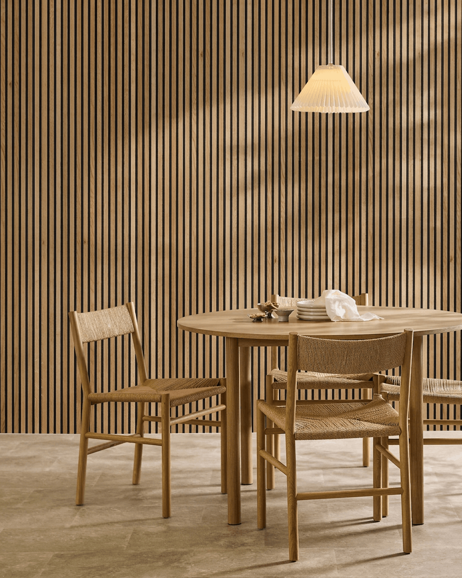 Minimalistyczna jadalnia z drewnianym stołem, dwoma krzesłami i akustycznym panelem drewnianym WoodUpp Akupanel w wykończeniu Rustic Brown Oak (300 x 60 cm), oświetlona wiszącą lampą.