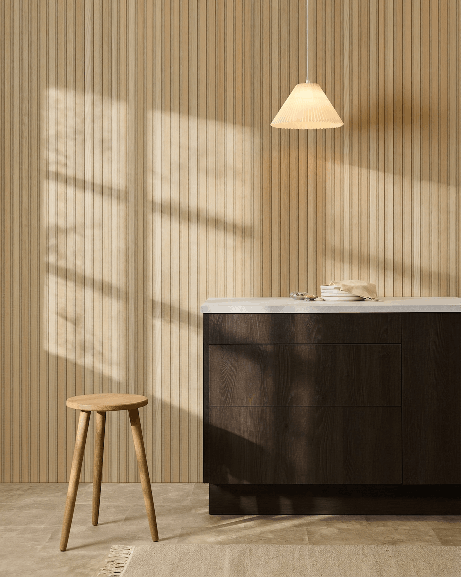 Skandynawska kuchnia z panelem akustycznym z lamelami WoodUpp Akupanel Classic Oak, ciemnymi drewnianymi szafkami, drewnianym stołkiem, lampą wiszącą i światłem słonecznym rzucającym cienie.