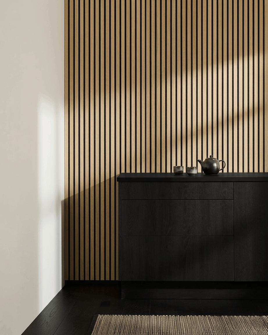 Minimalistyczna kuchnia w stylu nordyckim z WoodUpp Akupanel Panel akustyczny w wykończeniu Classic Oak, szafką black, czajnikiem, filiżankami i plecionym dywanikiem na ciemnej drewnianej podłodze.