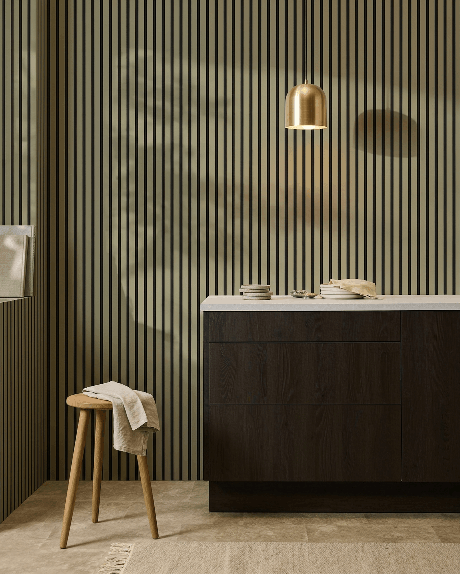 Nowoczesna kuchnia z ciemnymi drewnianymi szafkami, marmurowym blatem, brass lampą wiszącą oraz akustycznym panelem ściennym z listwami WoodUpp Akupanel Colored do pochłaniania dźwięku.