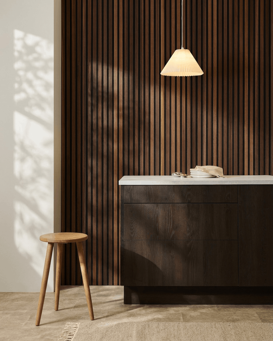 Minimalistyczne wnętrze z drewnianym stołkiem, szafką z ciemnego drewna, lampą wiszącą i akustycznym panel ściennym z drewnianych lameli WoodUpp Akupanel w kolorze Natural Wood/Smoked Larch.