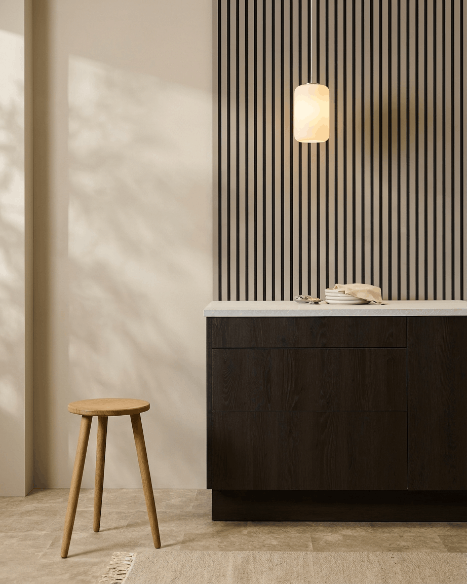 Minimalistyczna kuchnia z panelami akustycznymi WoodUpp Akupanel (Stone, Black Felt) na ścianie, drewnianym stołkiem, ciemnymi szafkami i lampą wiszącą w neutralnych tonach.
