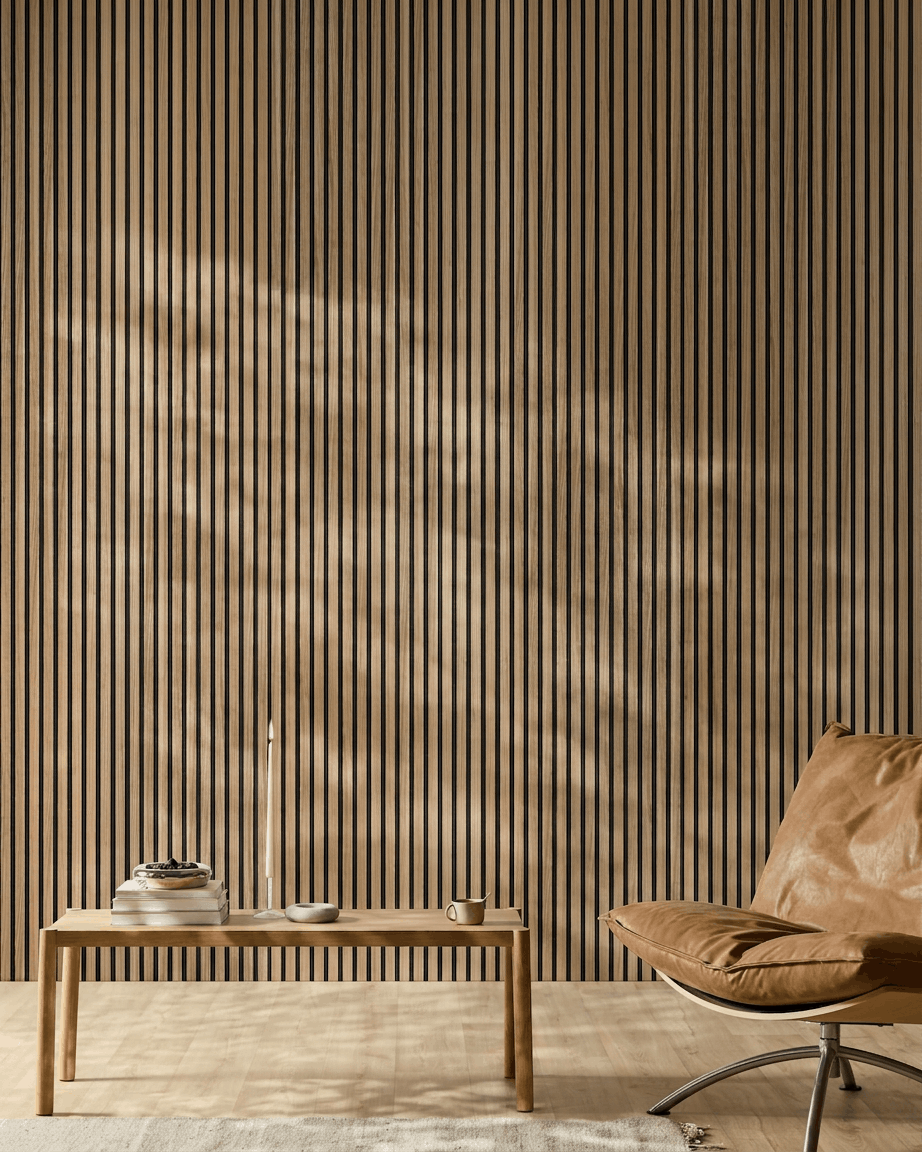 Minimalistyczne wnętrze z WoodUpp Akupanel Panel akustyczny (lamele English Walnut, filc black), skórzanym fotelem w kolorze tan, stołem z jasnego drewna, świecą, książkami, filiżanką i białym dywanem w słońcu.