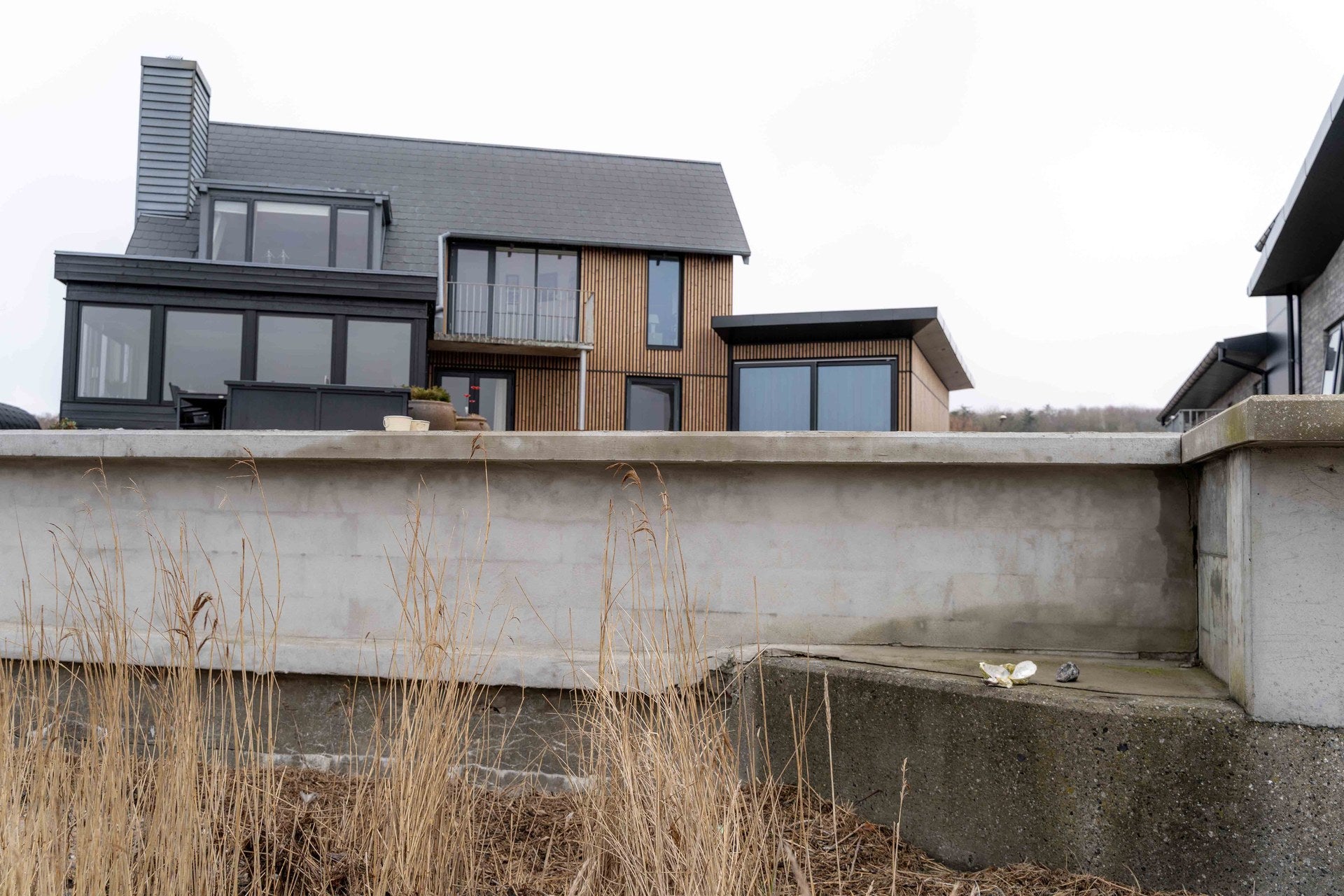 Lemvig house with brown Aluwood slat cladding and black window frames seen from the beach side over a concrete sea wall and reeds