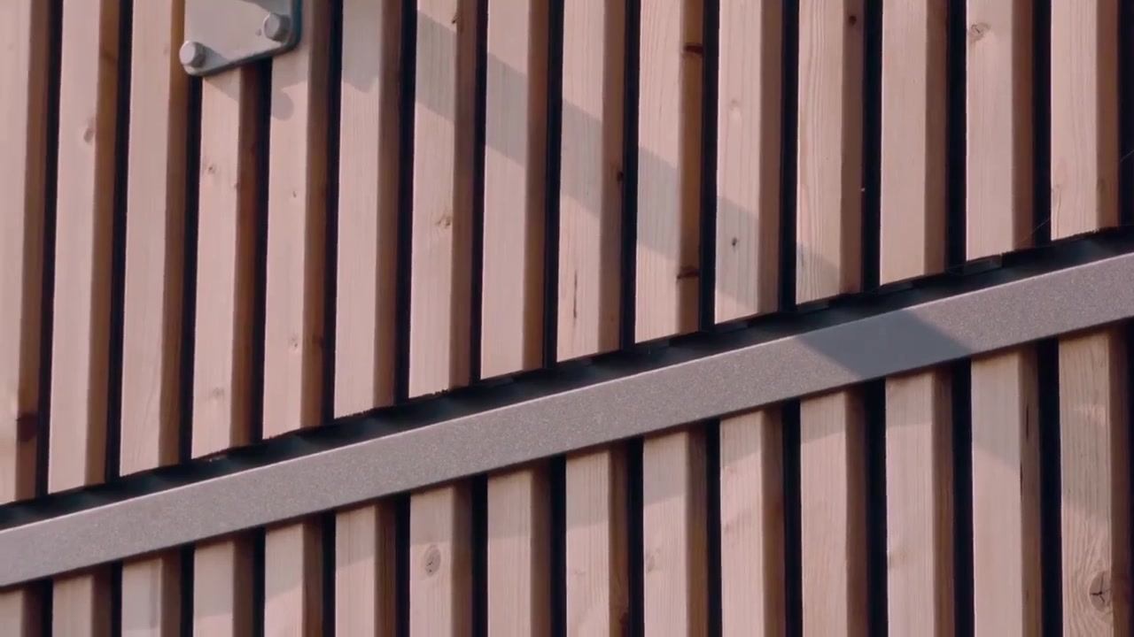 Side wall of a workshop building clad in vertical Aluwood slats with white windows, downpipe and gravel yard