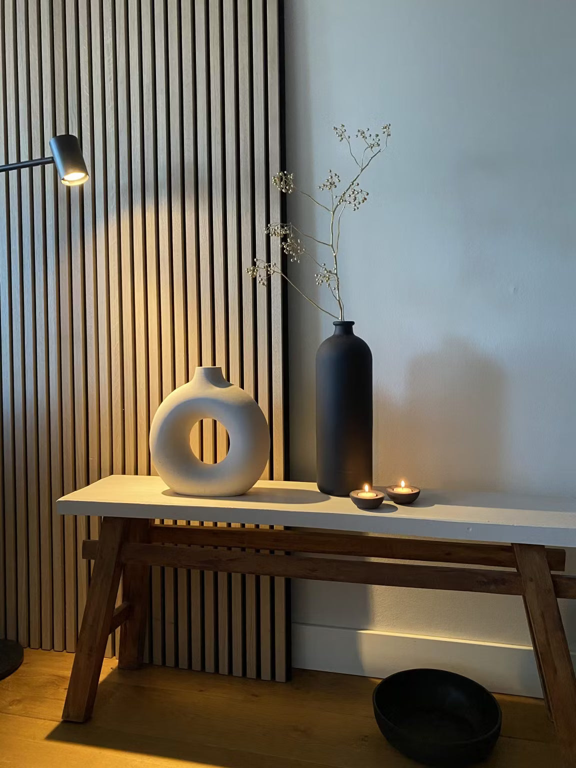 Minimalistyczny wystrój z białym okrągłym wazonem, czarnym wysokim wazonem z suszonymi łodygami, dwiema świeczkami tealight i czarną misą na tle panelu akustycznego z listew drewnianych Akupanel.
