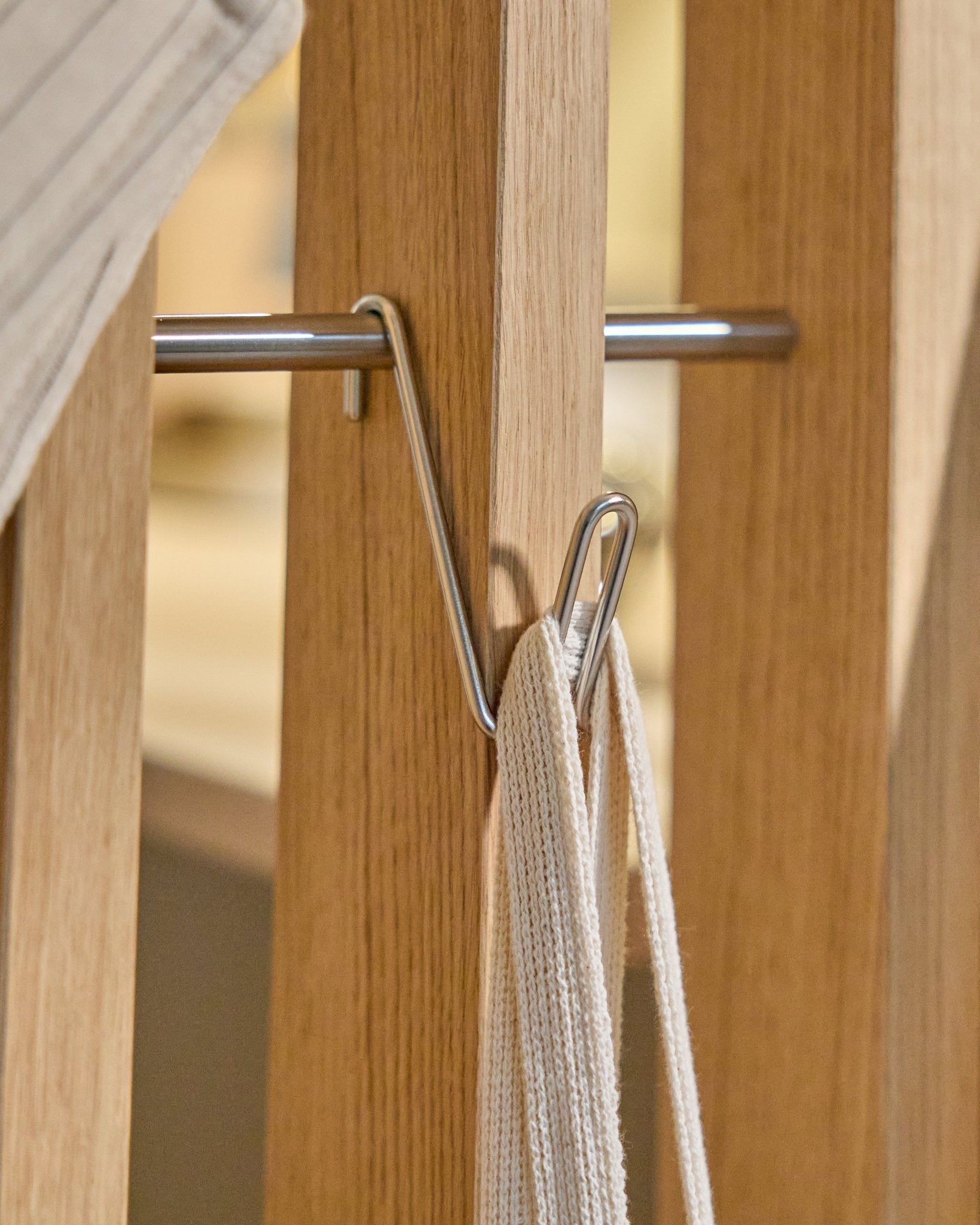 Haczyk Alva Wire Hook / BM1 (stal nierdzewna Steel, krótki) od WoodUpp jest zamontowany na drewnianej szynie Panel akustyczny i podtrzymuje biały pasek z metalowym prętem; tło beige.