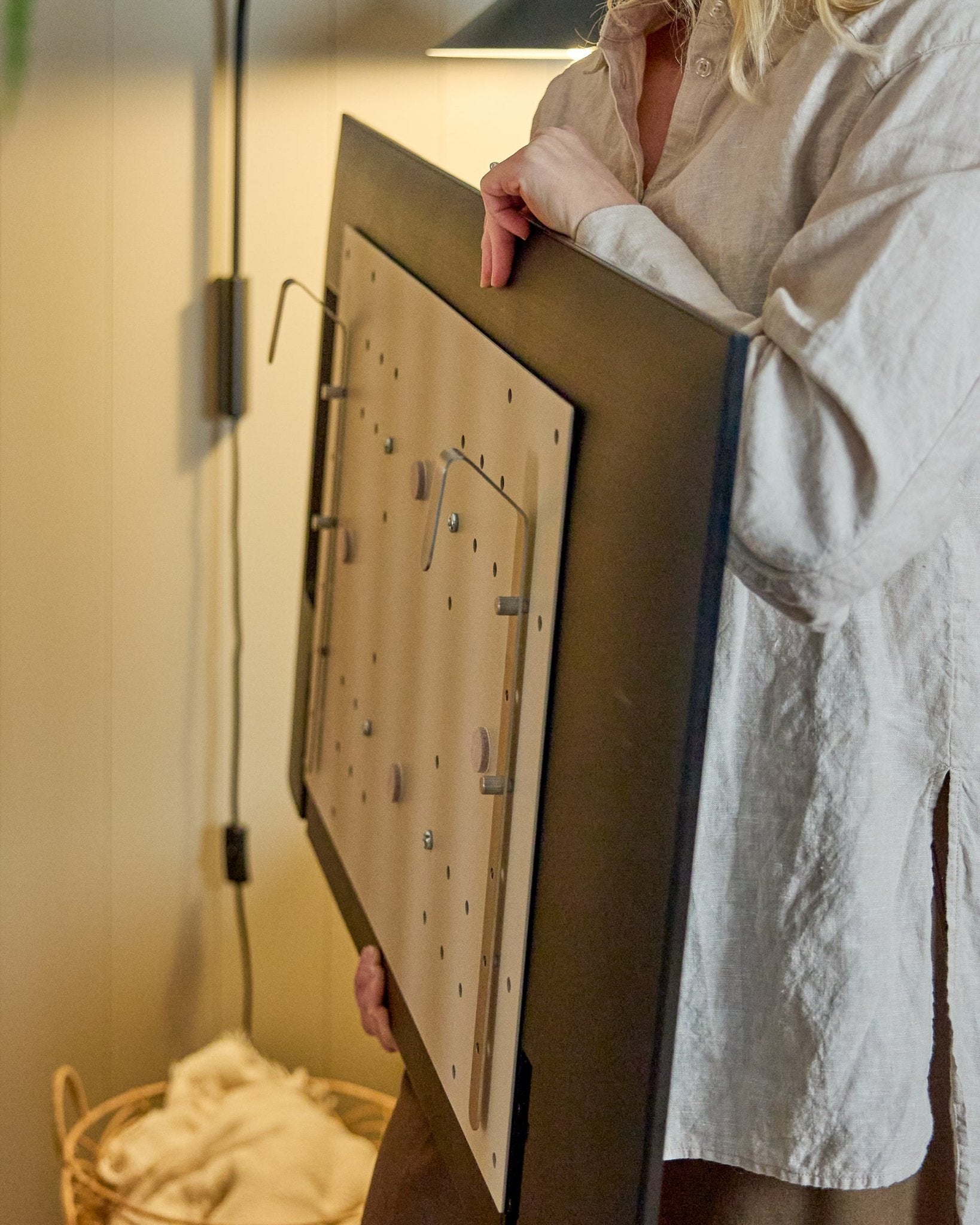 Osoba w jasnej koszuli trzyma WoodUpp Nell BM1 TV-Mount w kolorze beige, 64x44 cm — kompatybilny z nordyckimi akustycznymi panelami drewnianymi, do instalacji bez montażu na ścianie.