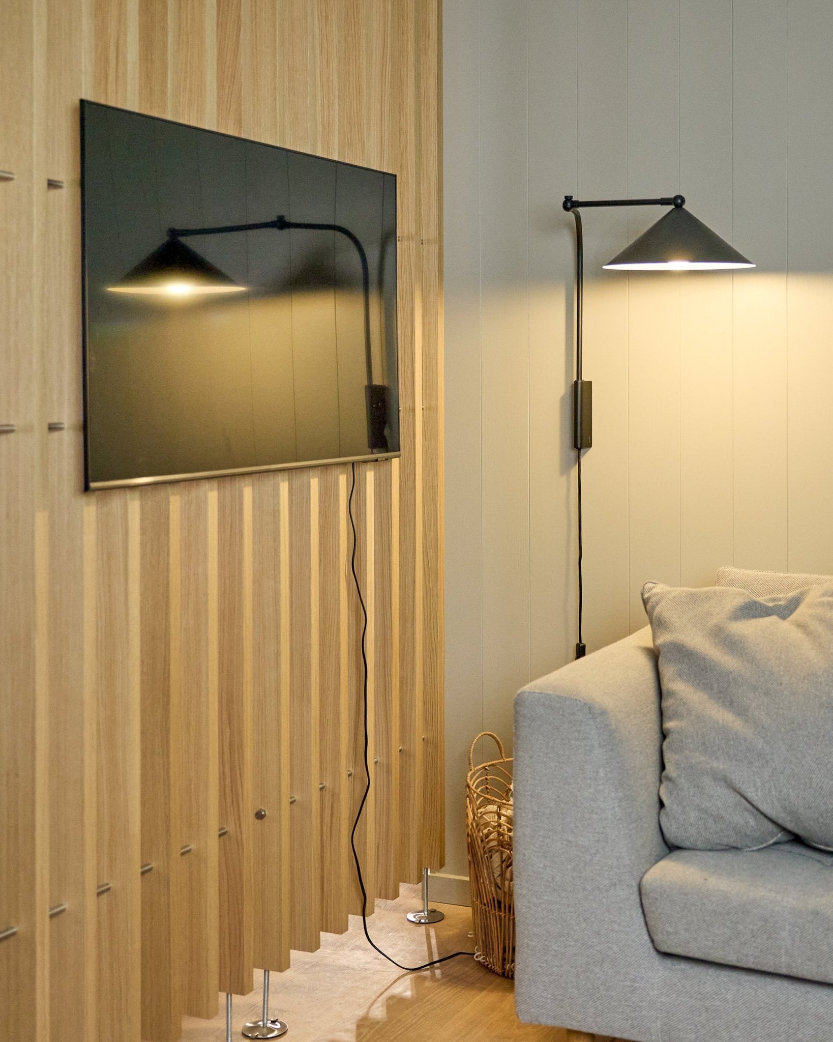Nell | TV-Mount / BM1 od WoodUpp (Beige, 64x44 cm) stabilnie mocuje telewizor z płaskim ekranem na nordyckim Panel akustyczny obok szarej sofy; w pobliżu stoi kosz.
