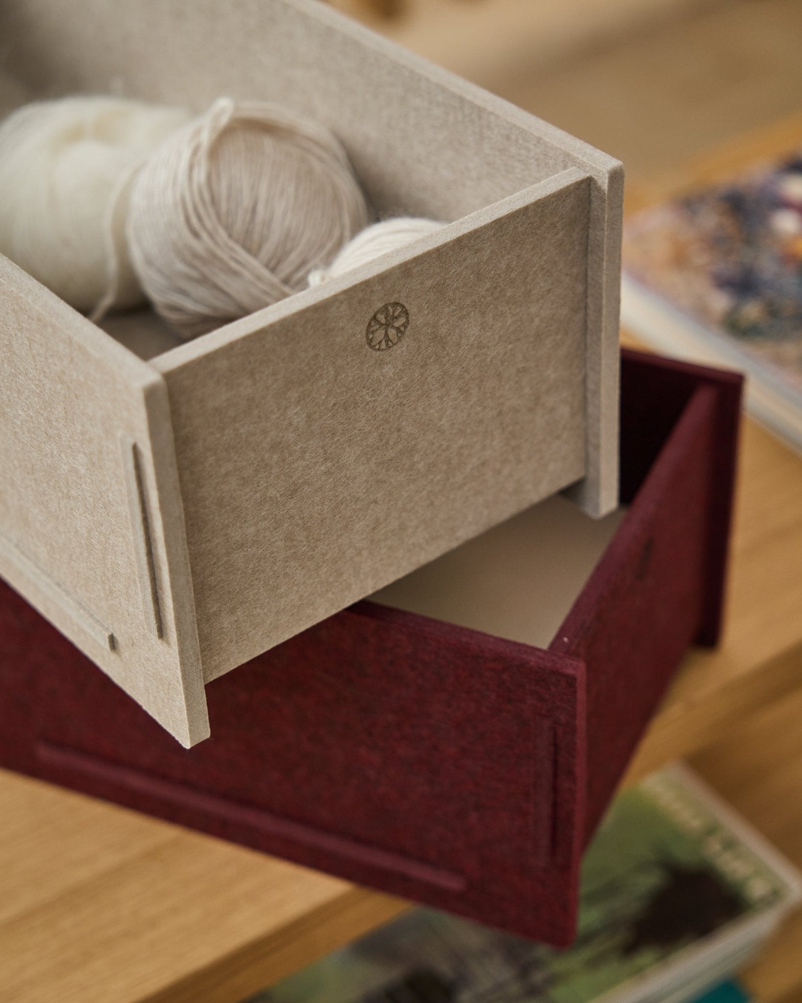beige pudełko do przechowywania z filcu PET wypełnione włóczką stoi na zamkniętym WoodUpp Vigg Felt Box/BM2 w kolorze ox red, ustawionym na drewnianej podłodze, z książkami w tle.