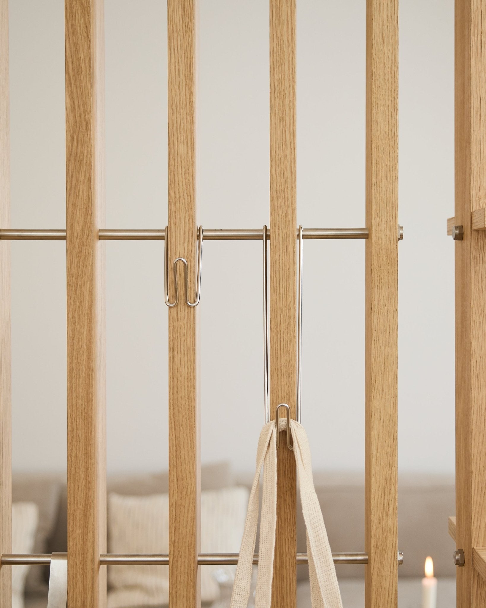 Zbliżenie na WoodUpp Alva | Wire Hook BM1 (stal nierdzewna steel, długi, zestaw 3 szt.) zamocowany do jasnych lameli Panel akustyczny z białym paskiem; sofa i świeca są rozmyte w tle.
