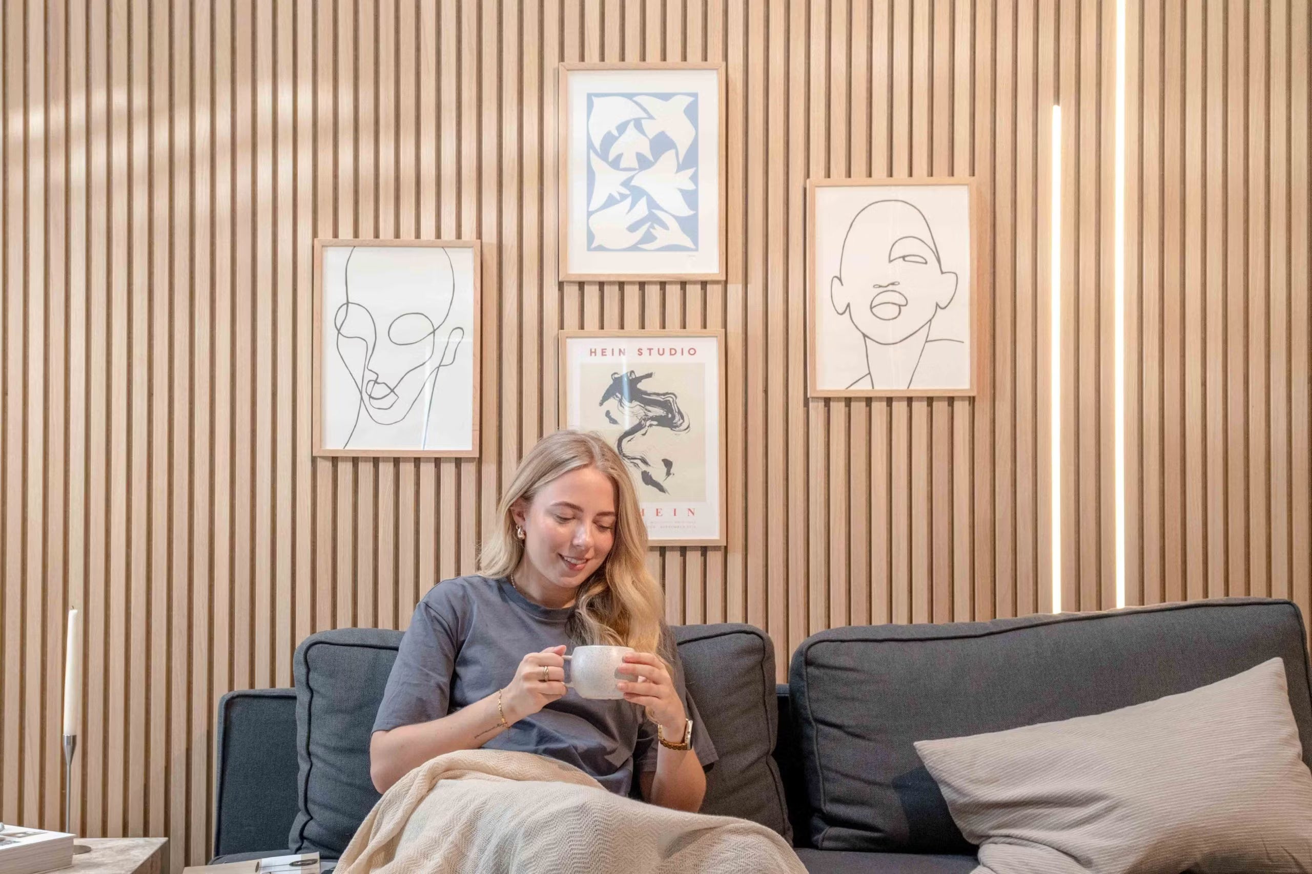 Kobieta relaksująca się na sofie z kubkiem, uśmiechnięta pod minimalistyczną grafiką liniową i abstrakcyjnym wystrojem zamontowanym na teksturowanych skandynawskich akustycznych panelach ściennych.